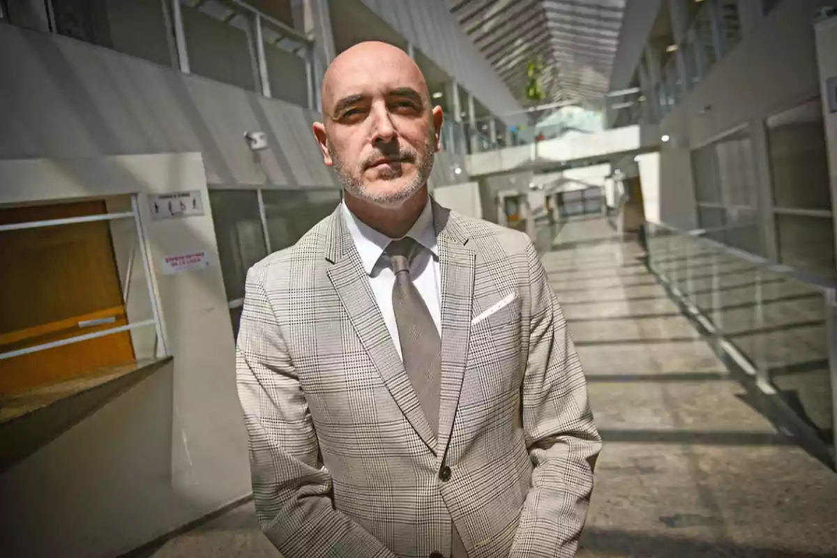 Hombre calvo con barba de pocos días usando traje claro y corbata posando en un pasillo moderno y luminoso de un edificio. Hombre calvo con barba de pocos días usando traje claro y corbata posando en un pasillo moderno y luminoso de un edificio.