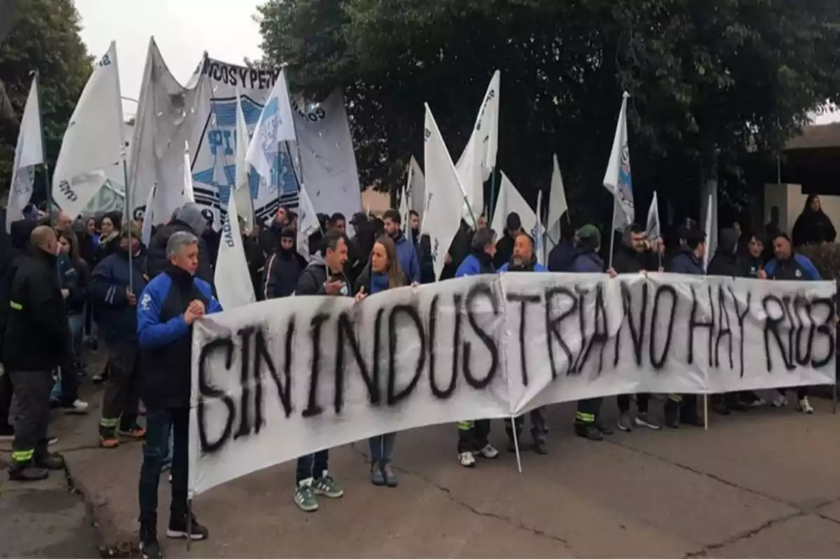 Un grupo de personas participa en una manifestación sosteniendo una pancarta blanca con letras negras que dice SIN INDUSTRIA NO HAY RIQUEZA y varias banderas blancas ondean detrás de ellos