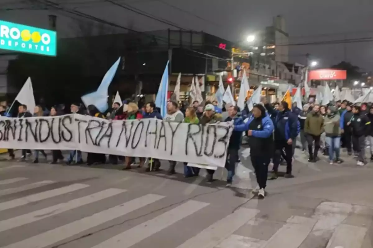 Manifestación nocturna en la calle con personas sosteniendo una pancarta que dice sin industria no hay ríos y varias banderas ondeando Manifestación nocturna en la calle con personas sosteniendo una pancarta que dice sin industria no hay ríos y varias banderas ondeando