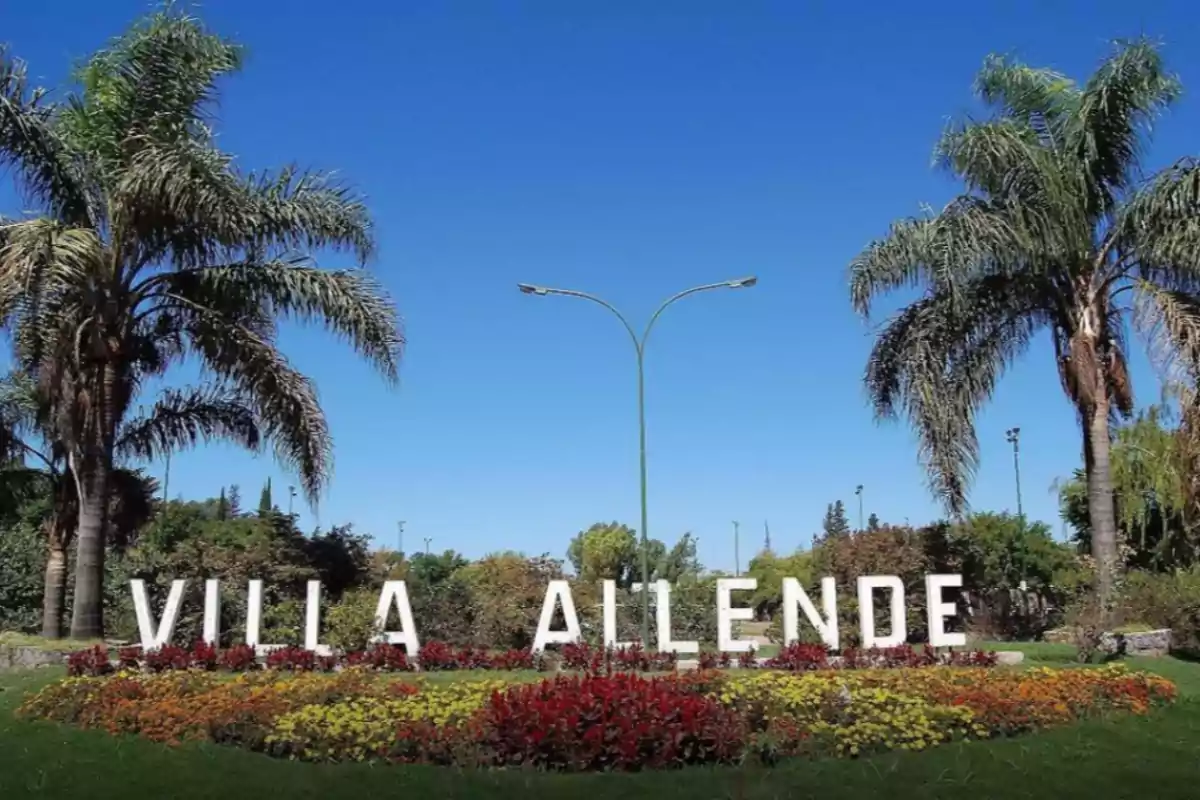 Letrero de Villa Allende rodeado de palmeras y flores coloridas bajo un cielo despejado.
