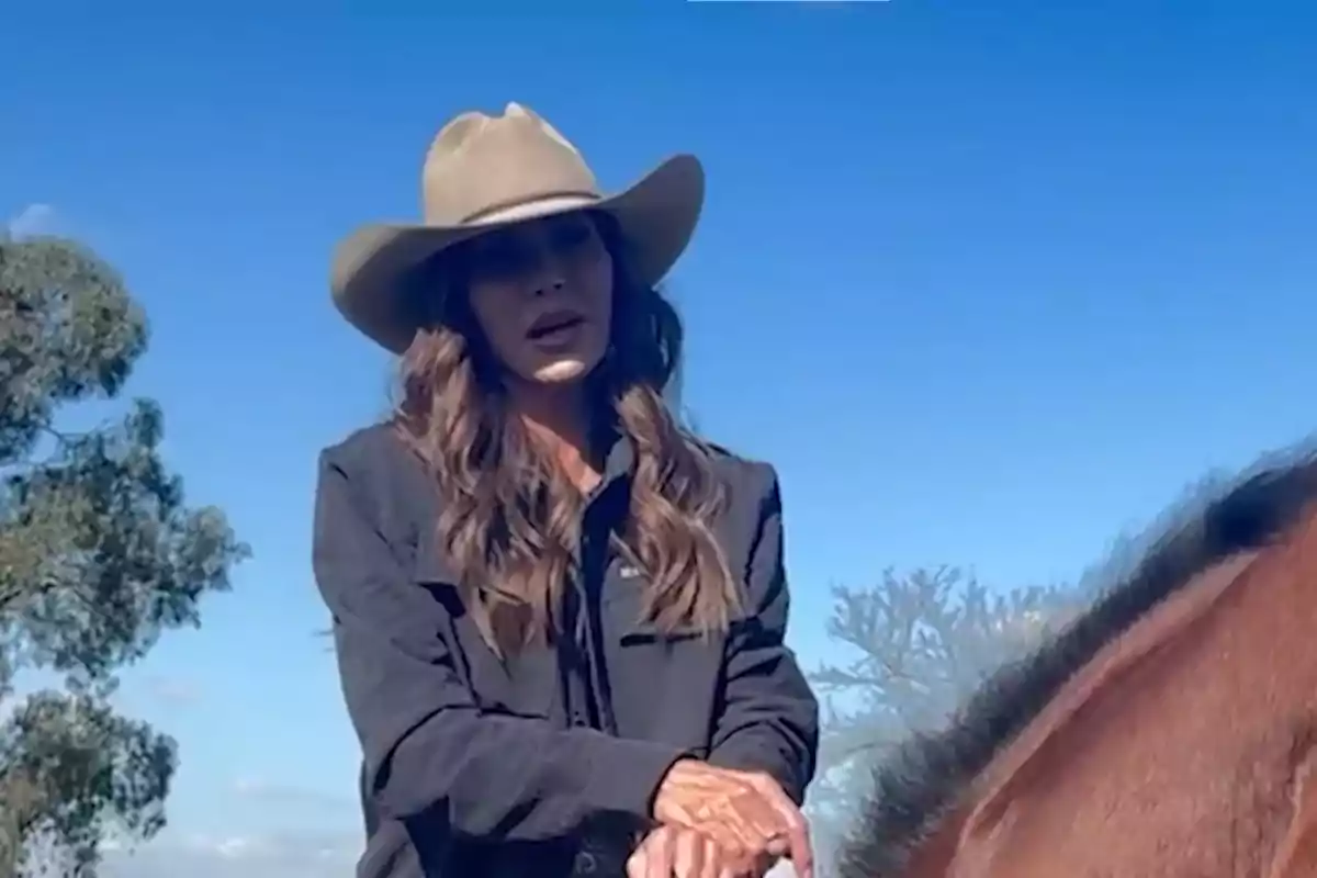 Mujer con sombrero vaquero y chaqueta oscura montando un caballo bajo un cielo azul