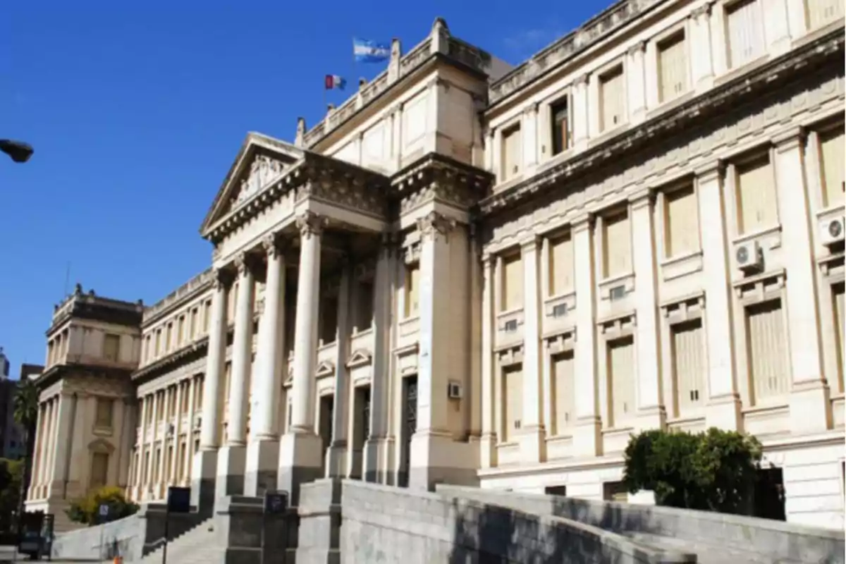Fachada de un edificio antiguo de estilo neoclásico con columnas y banderas en el techo bajo un cielo despejado Fachada de un edificio antiguo de estilo neoclásico con columnas y banderas en el techo bajo un cielo despejado