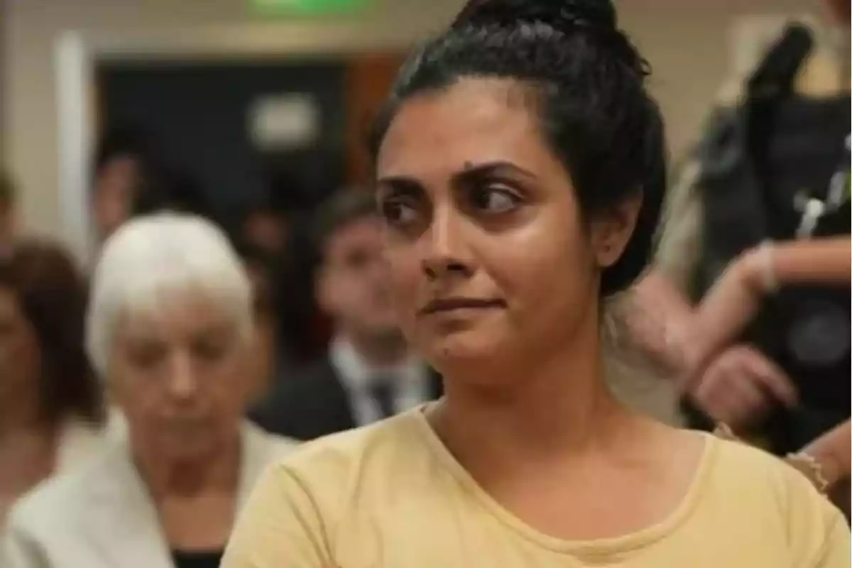 Mujer de cabello oscuro recogido con expresión seria en un lugar público rodeada de otras personas desenfocadas al fondo