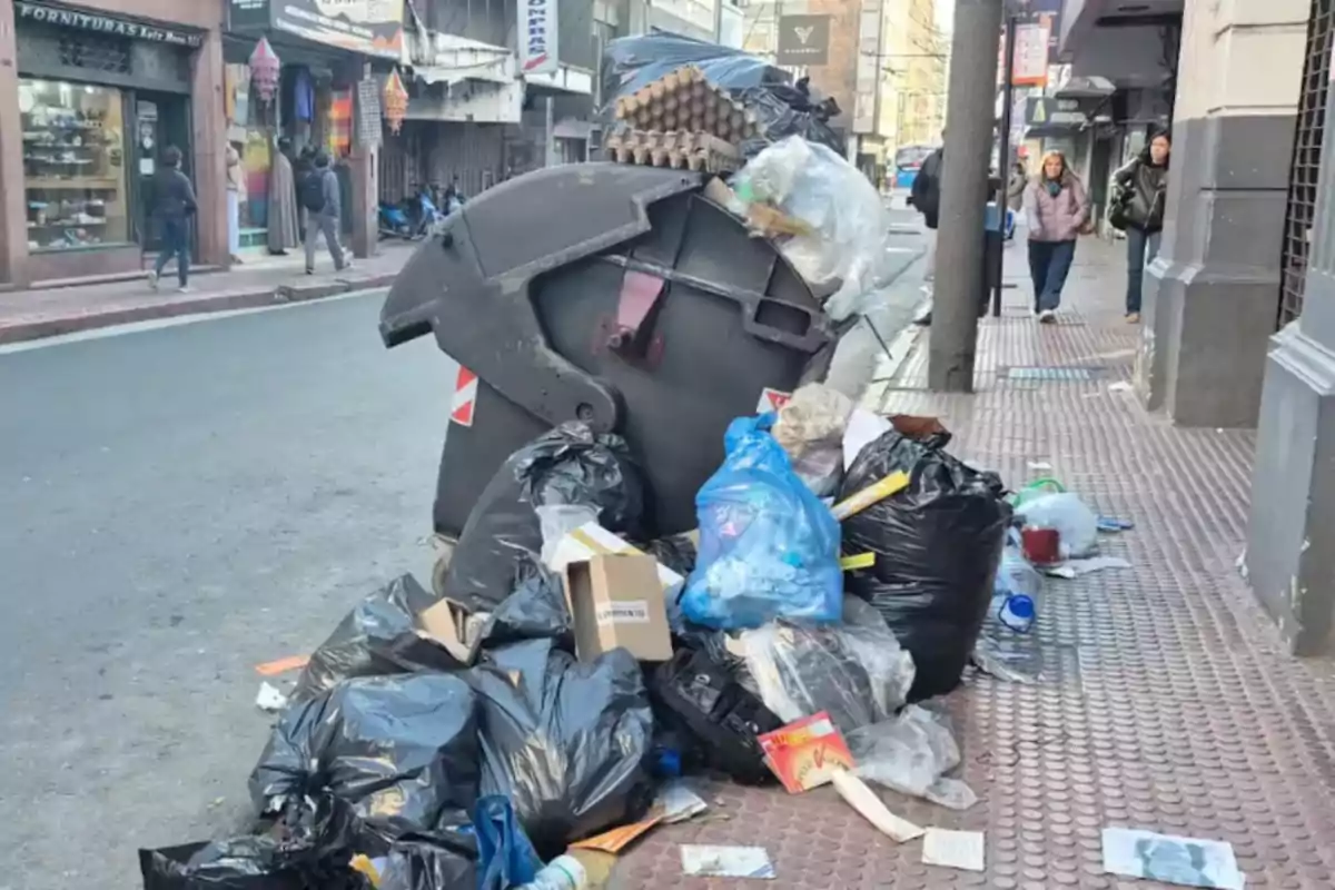 Contenedor de basura desbordado con bolsas y residuos acumulados en la vereda de una calle urbana Contenedor de basura desbordado con bolsas y residuos acumulados en la vereda de una calle urbana