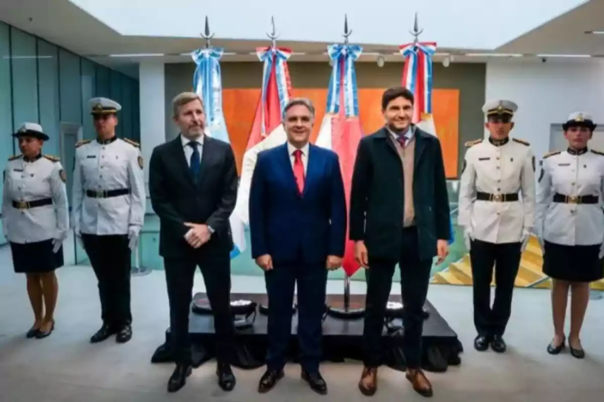 Tres hombres de traje posan al centro junto a seis personas uniformadas en un ambiente formal con banderas y decoraciones patrias al fondo Tres hombres de traje posan al centro junto a seis personas uniformadas en un ambiente formal con banderas y decoraciones patrias al fondo