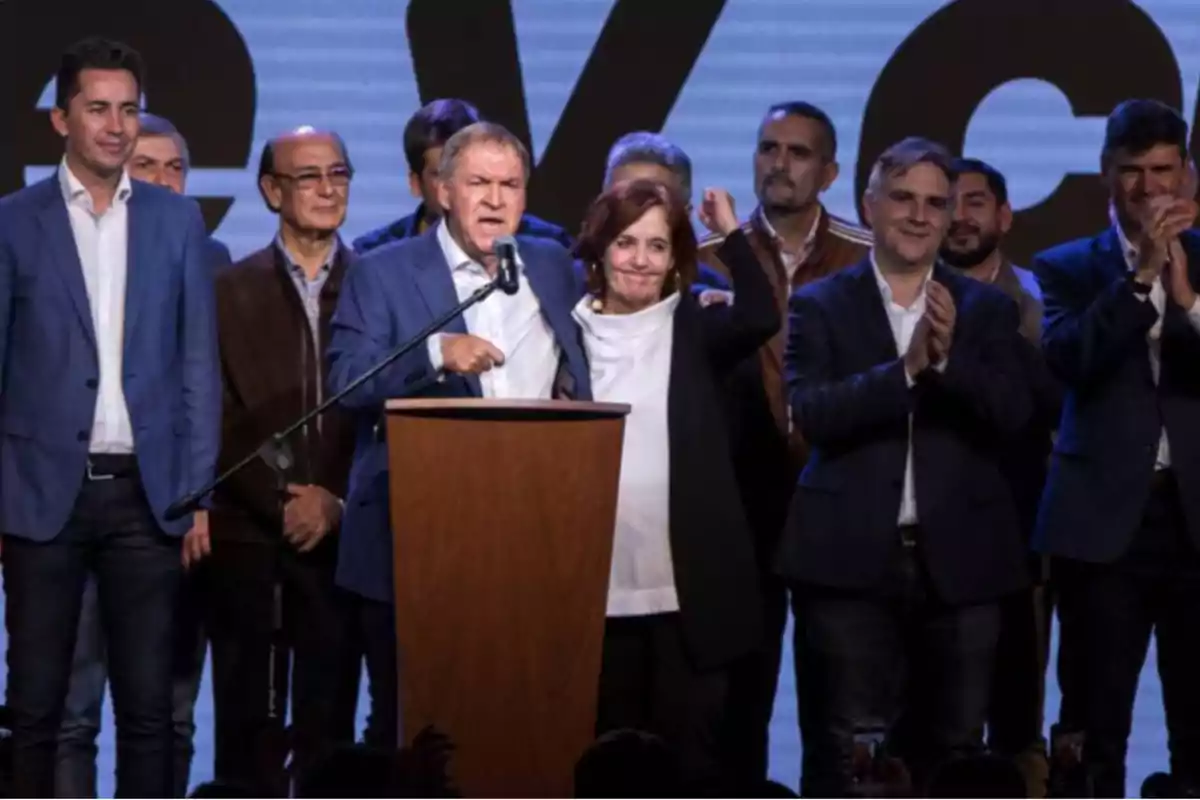 A group of people dressed formally on a stage while one of them speaks into a microphone and the others applaud.