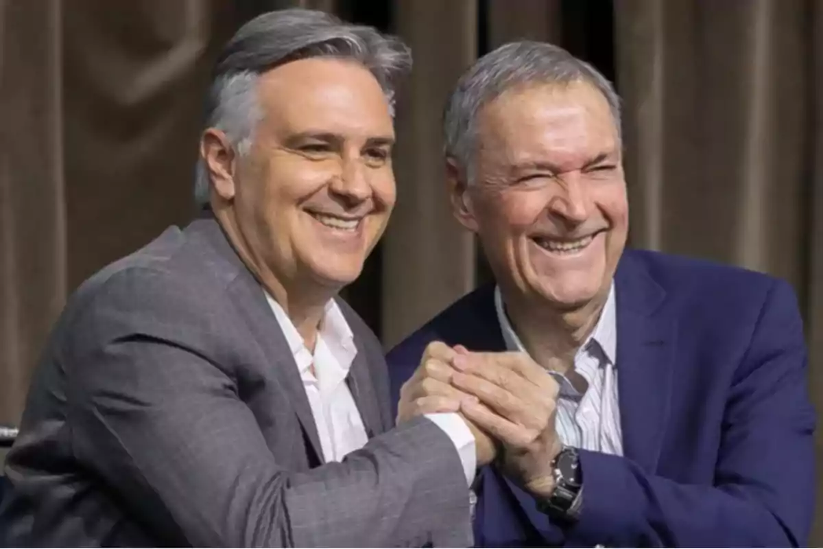 Dos hombres sonrientes se dan la mano y posan juntos usando trajes formales Dos hombres sonrientes se dan la mano y posan juntos usando trajes formales