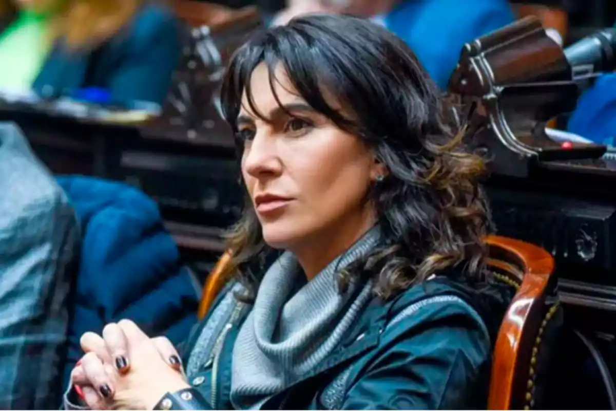 Mujer de cabello oscuro sentada en lo que parece ser una sala de reuniones o parlamento con expresión seria y las manos entrelazadas. Mujer de cabello oscuro sentada en lo que parece ser una sala de reuniones o parlamento con expresión seria y las manos entrelazadas.