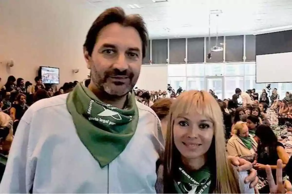 Un hombre y una mujer posan juntos en primer plano dentro de un salón lleno de personas, ambos llevan pañuelos verdes en el cuello y al fondo se observa un ambiente de reunión o asamblea. Un hombre y una mujer posan juntos en primer plano dentro de un salón lleno de personas, ambos llevan pañuelos verdes en el cuello y al fondo se observa un ambiente de reunión o asamblea.