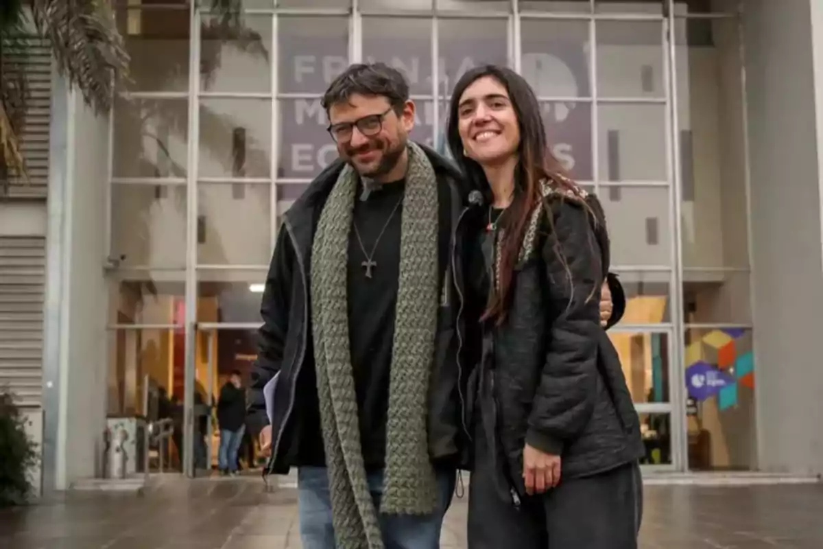 Dos personas sonríen y posan abrazadas frente a un edificio de vidrio en un día nublado. Dos personas sonríen y posan abrazadas frente a un edificio de vidrio en un día nublado.