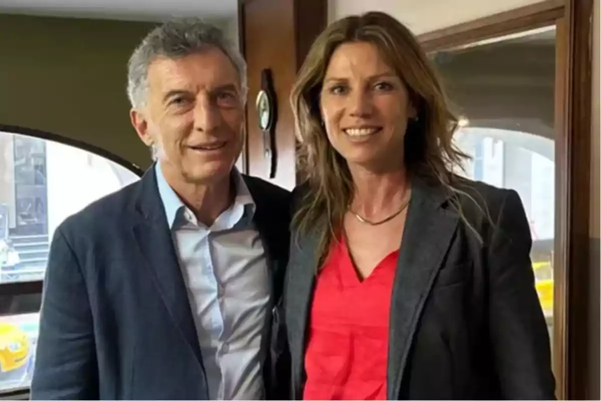 A man and a woman smile together indoors with windows in the background.