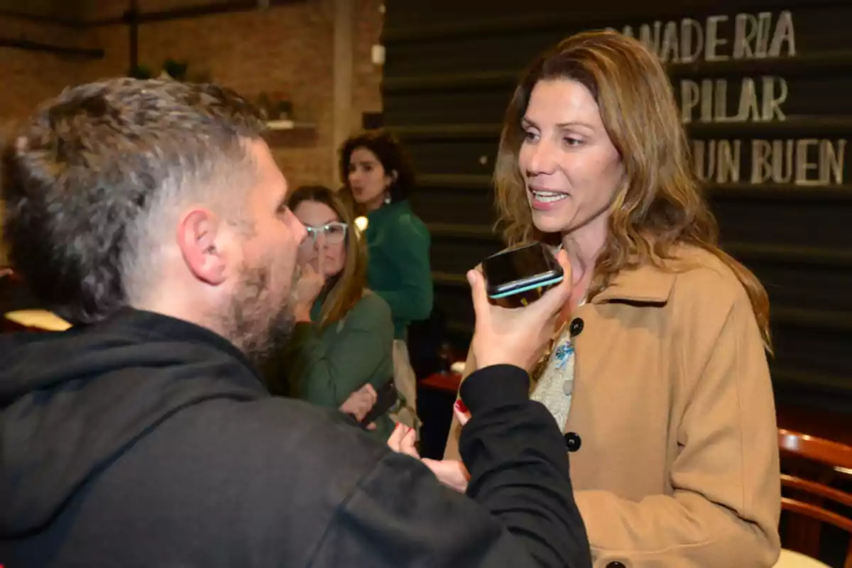 A man is holding a mobile phone while interviewing a light brown-haired woman wearing a beige coat; in the background, there are other people and a sign on the wall. A man is holding a mobile phone while interviewing a light brown-haired woman wearing a beige coat; in the background, there are other people and a sign on the wall.