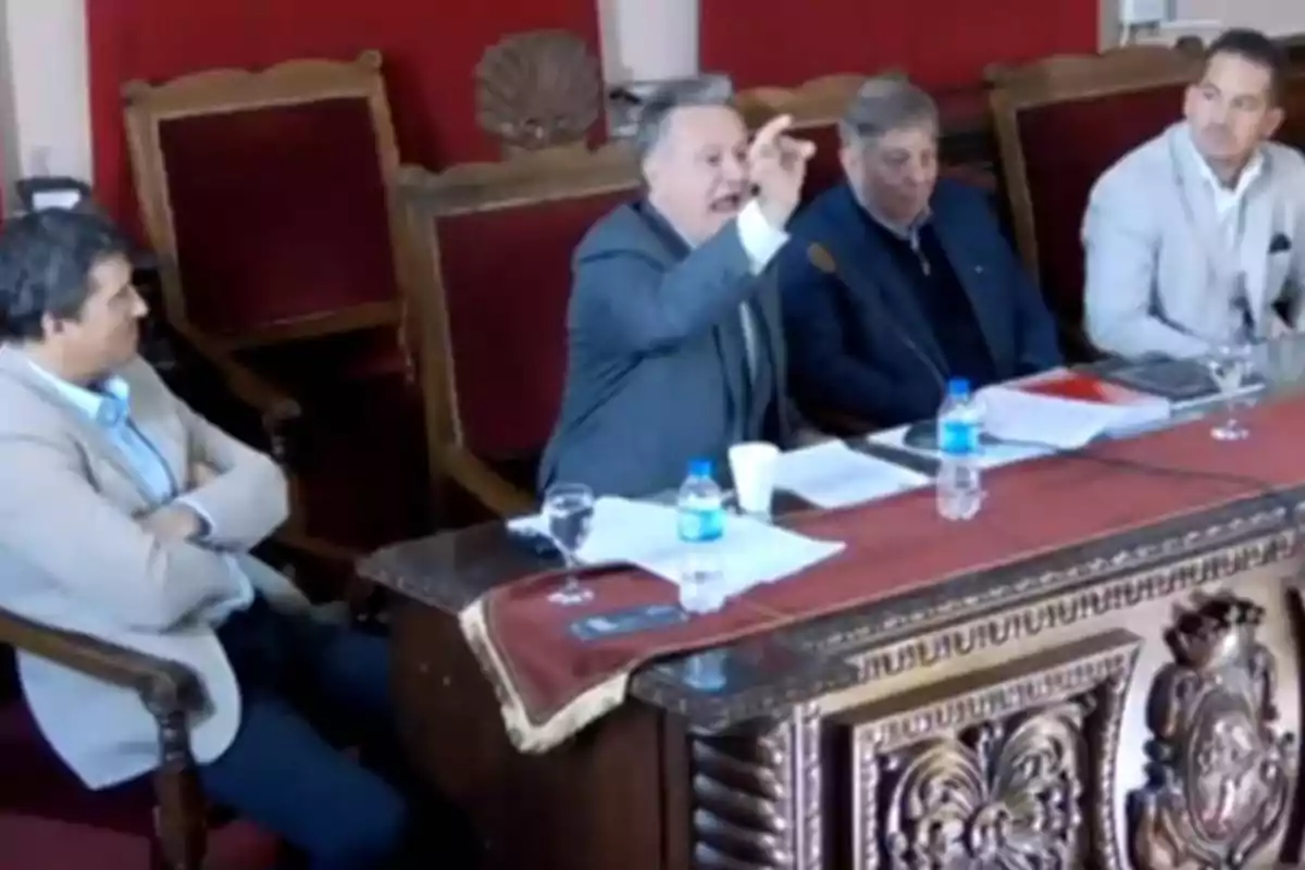 Four men in suits are sitting at a carved wooden table; one of them is gesturing while speaking, and there are water bottles, papers, and glasses on the table. Four men in suits are sitting at a carved wooden table; one of them is gesturing while speaking, and there are water bottles, papers, and glasses on the table.