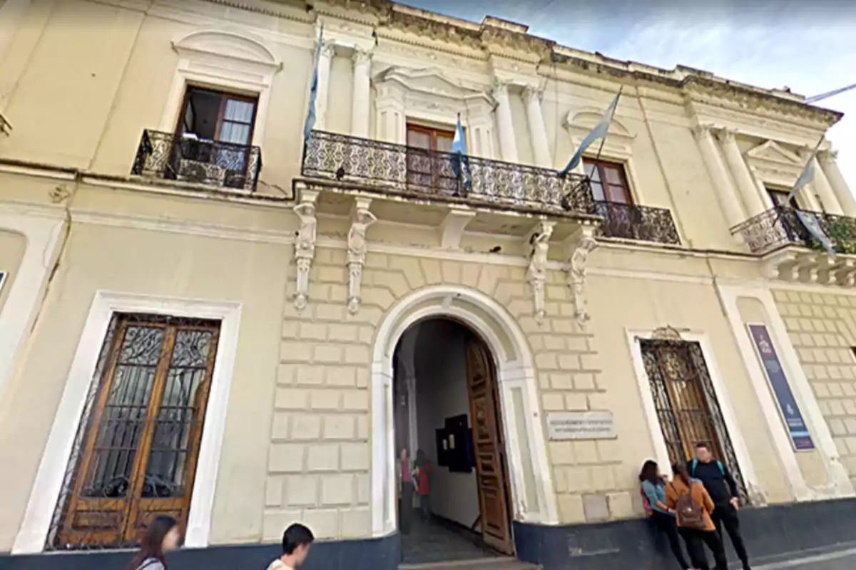 Fachada de un edificio antiguo de dos pisos con balcones de hierro y banderas argentinas, personas caminando y conversando en la vereda frente a la entrada principal. Fachada de un edificio antiguo de dos pisos con balcones de hierro y banderas argentinas, personas caminando y conversando en la vereda frente a la entrada principal.