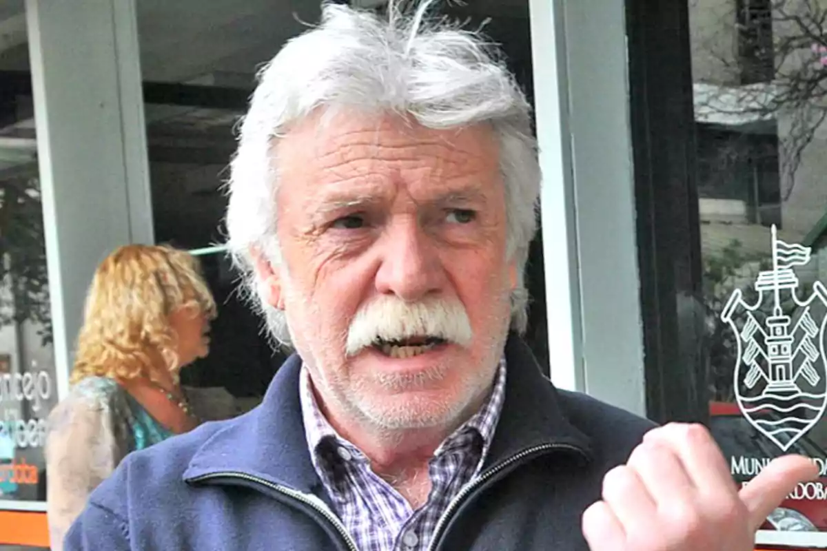 Hombre de cabello y bigote canoso con expresión seria señalando hacia un costado, al fondo una mujer desenfocada y una ventana con un logo blanco Hombre de cabello y bigote canoso con expresión seria señalando hacia un costado, al fondo una mujer desenfocada y una ventana con un logo blanco