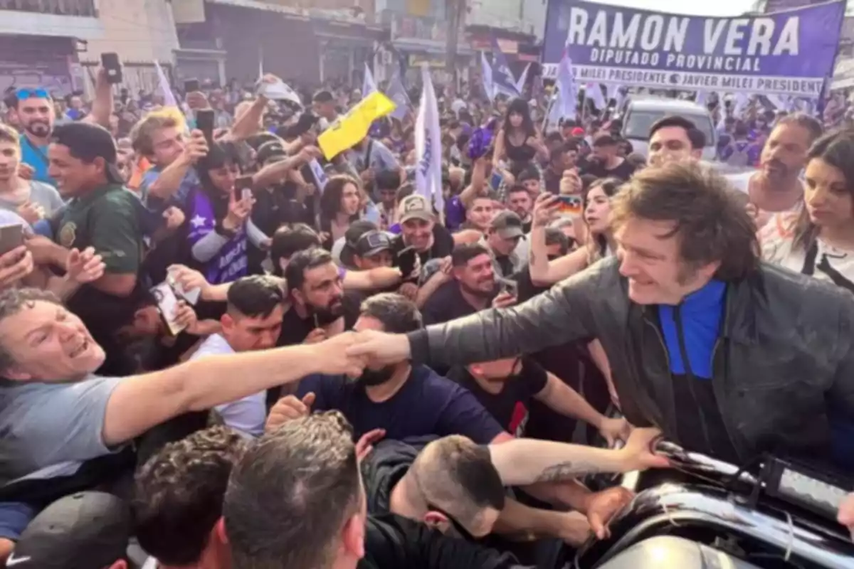 Un hombre sonriente saluda a una multitud entusiasta en un evento político al aire libre mientras muchas personas lo fotografían y sostienen banderas Un hombre sonriente saluda a una multitud entusiasta en un evento político al aire libre mientras muchas personas lo fotografían y sostienen banderas