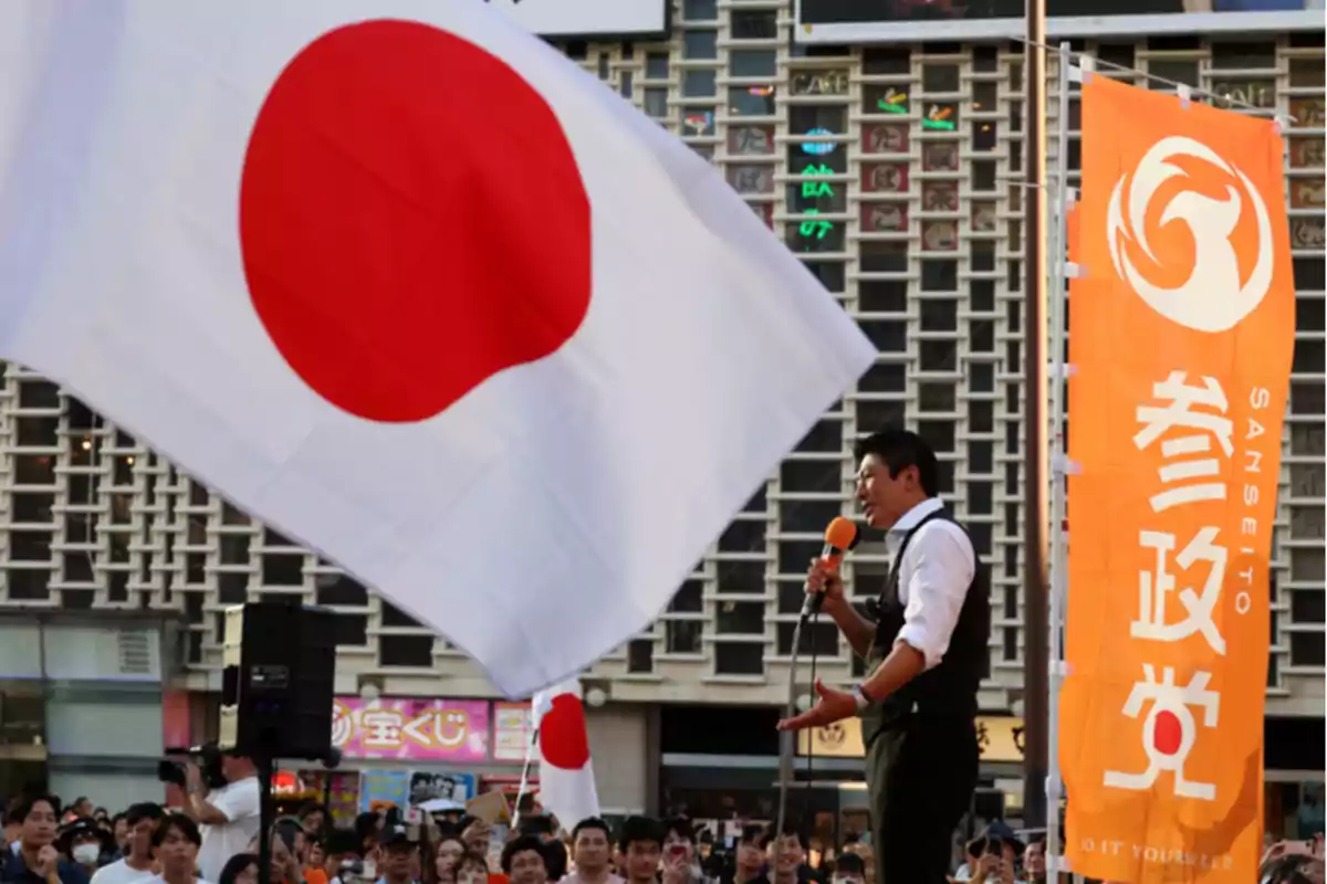 Un hombre habla por micrófono frente a una multitud junto a una gran bandera de Japón y una bandera naranja del partido Sanseito en un evento al aire libre Un hombre habla por micrófono frente a una multitud junto a una gran bandera de Japón y una bandera naranja del partido Sanseito en un evento al aire libre