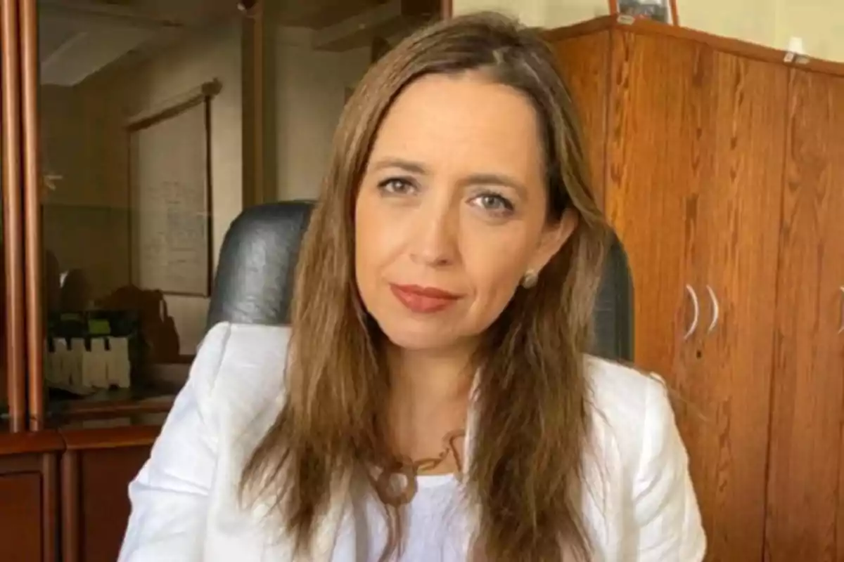 Long, straight brown-haired woman wearing a white blazer sitting in an office with wooden furniture in the background Long, straight brown-haired woman wearing a white blazer sitting in an office with wooden furniture in the background