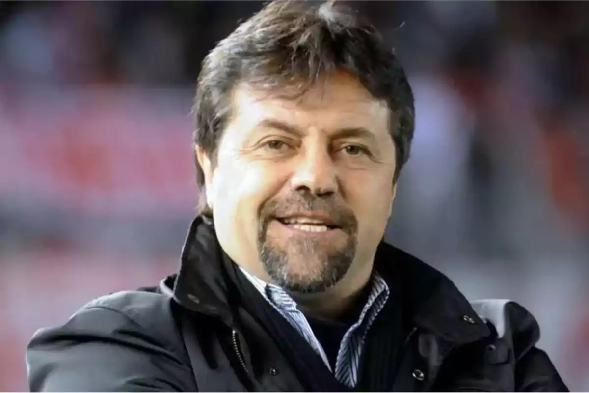 Hombre de mediana edad con barba y bigote, cabello castaño, vistiendo chaqueta oscura y camisa, sonriendo frente a un fondo desenfocado.