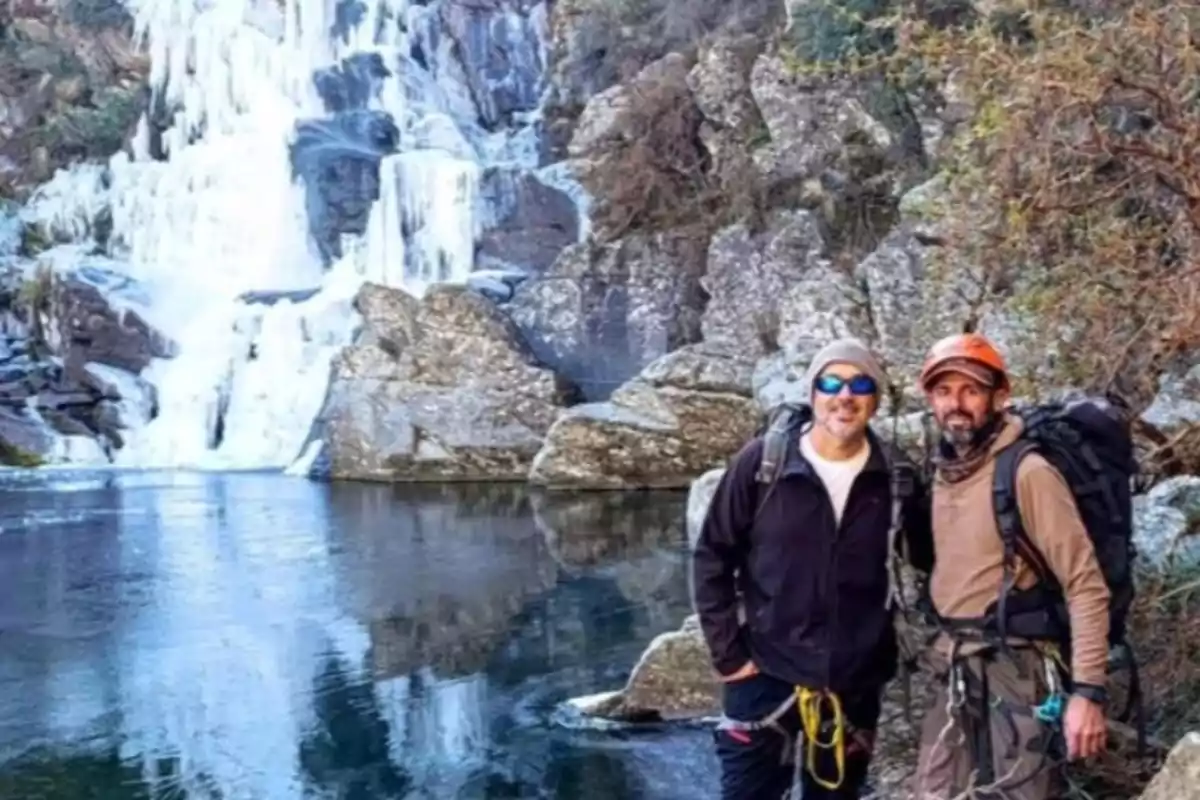 Dos hombres con equipo de senderismo posan frente a una cascada parcialmente congelada rodeada de rocas y vegetación
