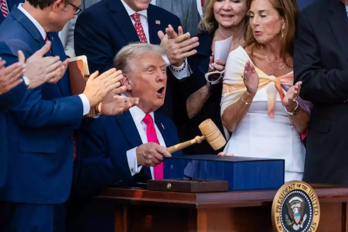 Un hombre de traje azul sostiene un mazo de madera mientras varias personas a su alrededor lo aplauden y observan con expresiones de entusiasmo junto a un escritorio con el sello presidencial de Estados Unidos.