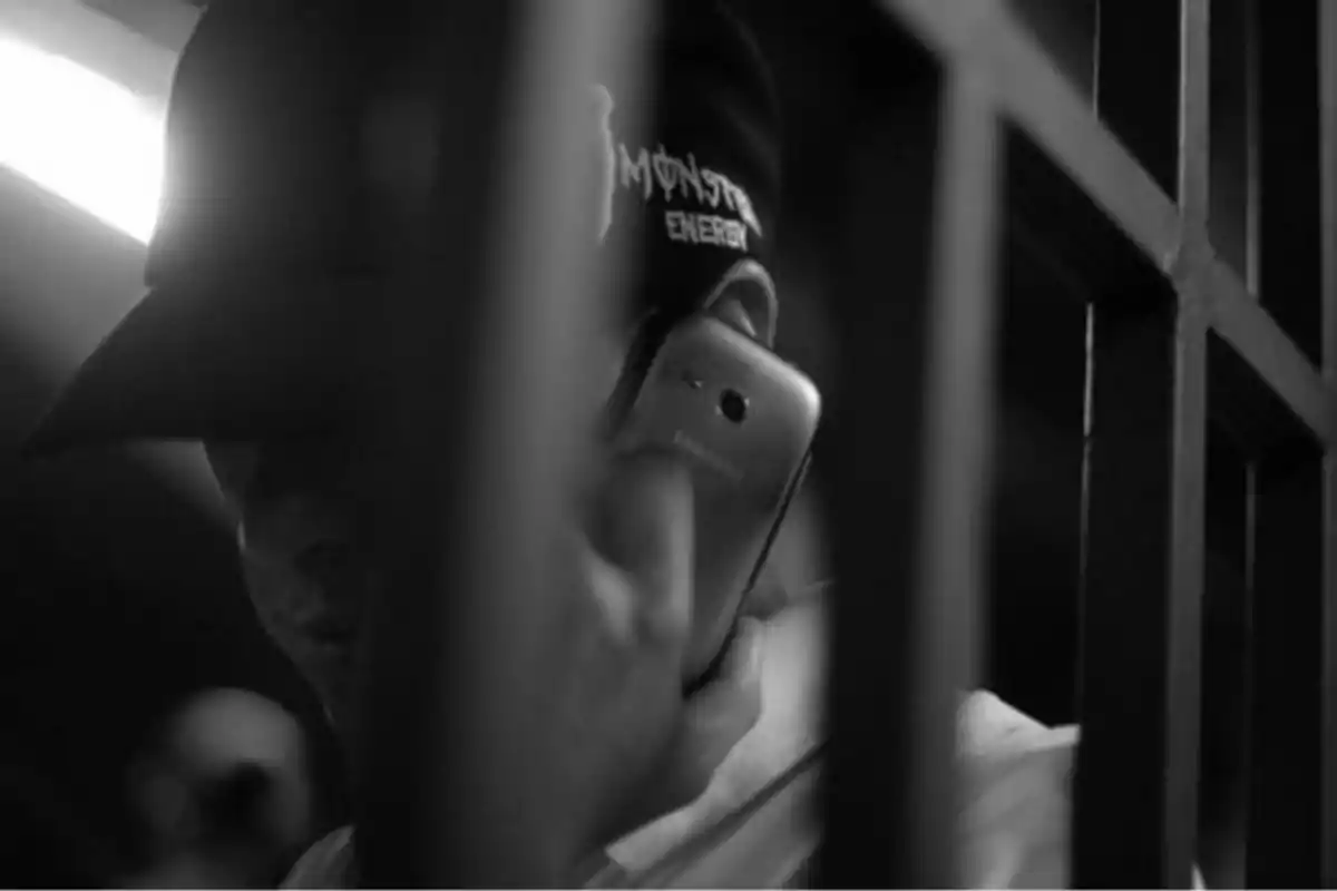 Persona con gorra hablando por teléfono móvil detrás de unos barrotes en una imagen en blanco y negro Persona con gorra hablando por teléfono móvil detrás de unos barrotes en una imagen en blanco y negro