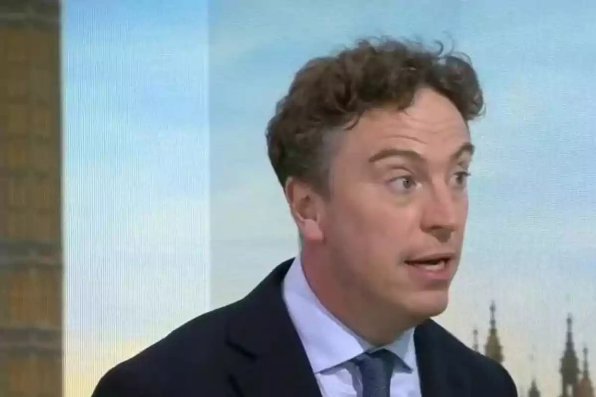 Curly-haired man in a dark suit speaking in a television studio with a backdrop of landmark buildings Curly-haired man in a dark suit speaking in a television studio with a backdrop of landmark buildings