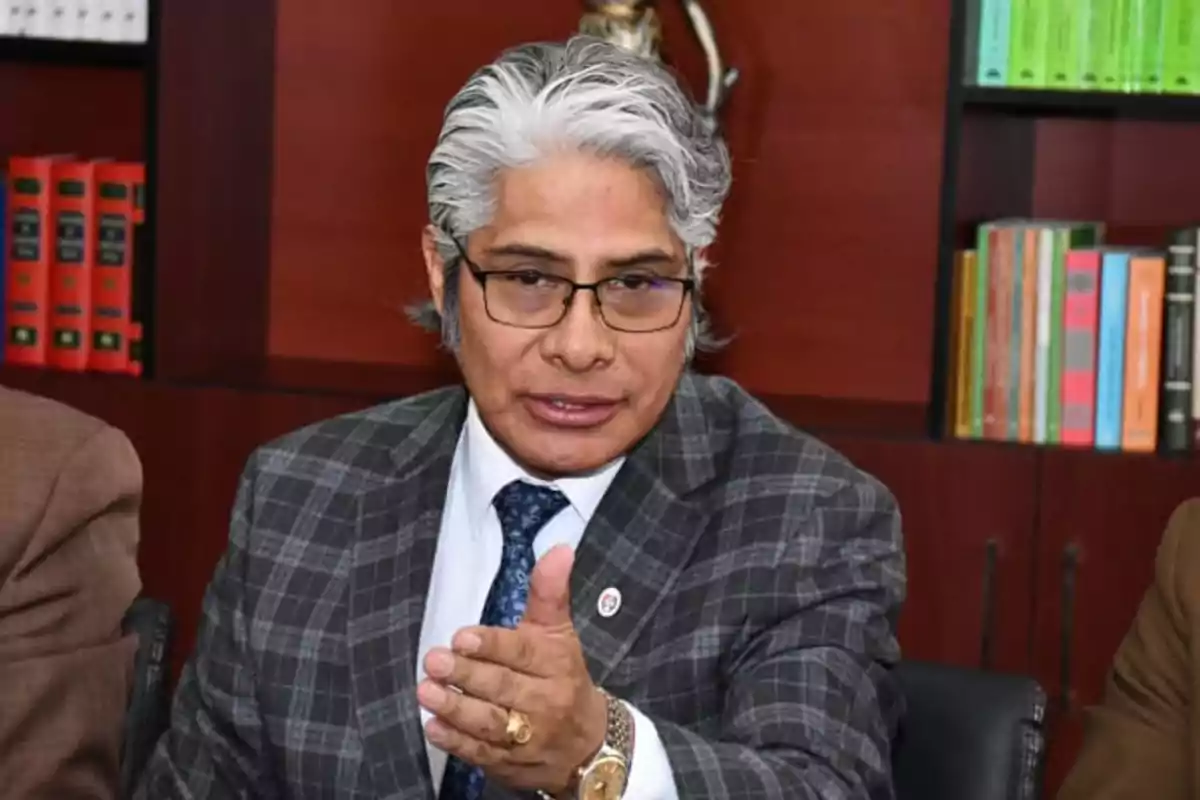 Hombre de cabello canoso y gafas con traje a cuadros sentado en una oficina con estanterías de libros al fondo mientras extiende la mano hacia el frente