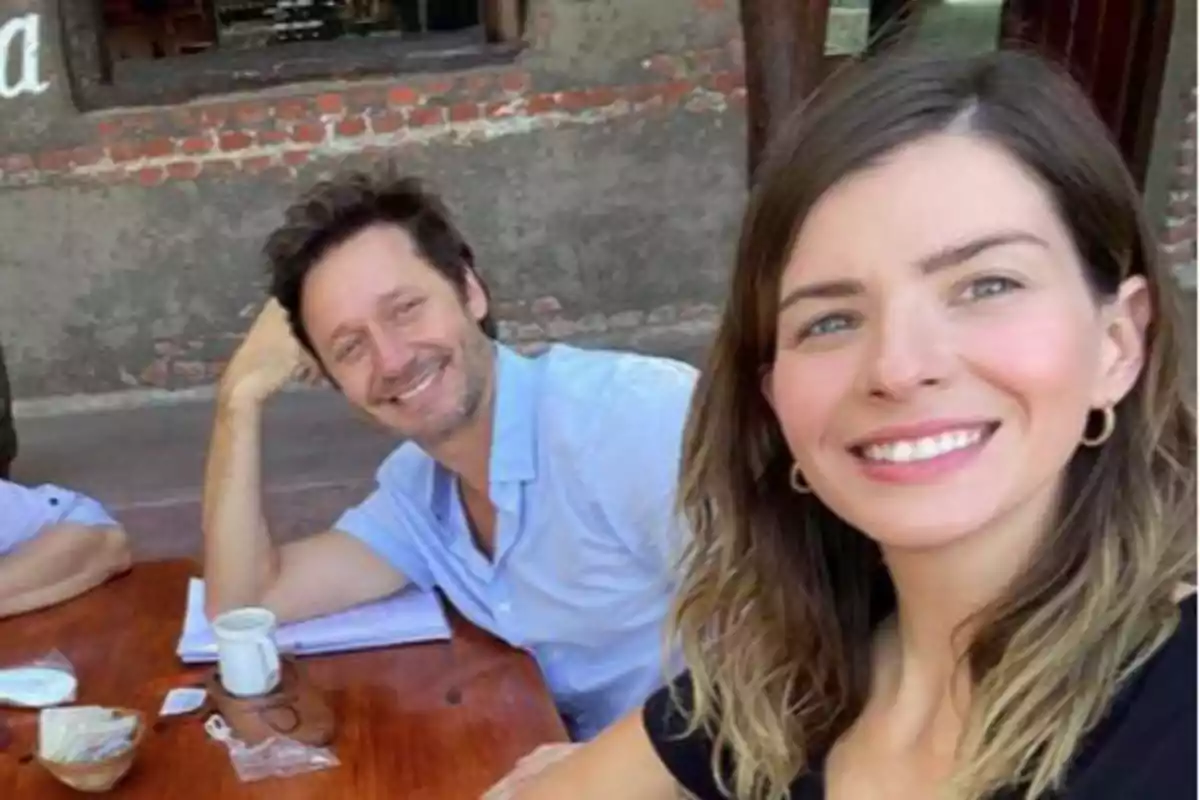 Dos personas sonríen mientras se toman una selfie sentados en una mesa de madera al aire libre con bebidas y comida sobre la mesa. Dos personas sonríen mientras se toman una selfie sentados en una mesa de madera al aire libre con bebidas y comida sobre la mesa.