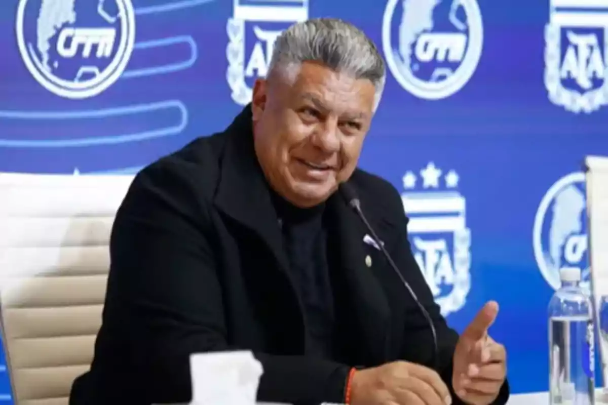 Hombre de cabello canoso hablando en una conferencia de prensa con fondo azul y logotipos deportivos Hombre de cabello canoso hablando en una conferencia de prensa con fondo azul y logotipos deportivos
