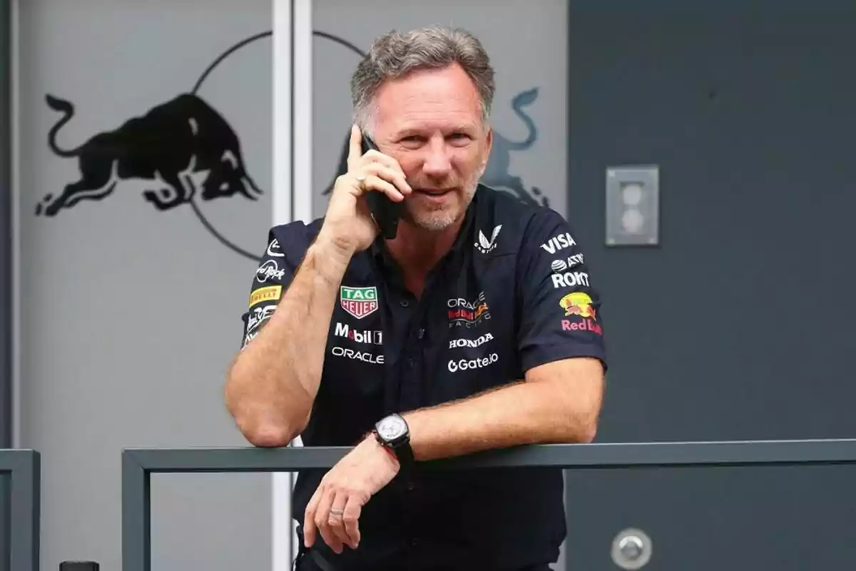 Hombre con camisa de Red Bull Racing hablando por teléfono móvil apoyado en una barandilla frente a un logo de toro. Hombre con camisa de Red Bull Racing hablando por teléfono móvil apoyado en una barandilla frente a un logo de toro.