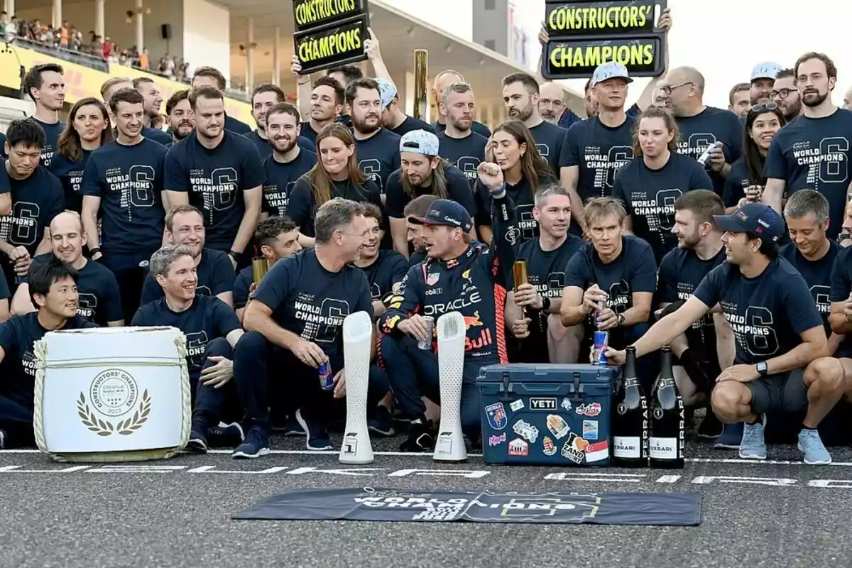 A large group of people in blue jerseys celebrate a motorsport constructors’ championship, holding trophies and champion signs while smiling and posing on a racetrack. A large group of people in blue jerseys celebrate a motorsport constructors’ championship, holding trophies and champion signs while smiling and posing on a racetrack.
