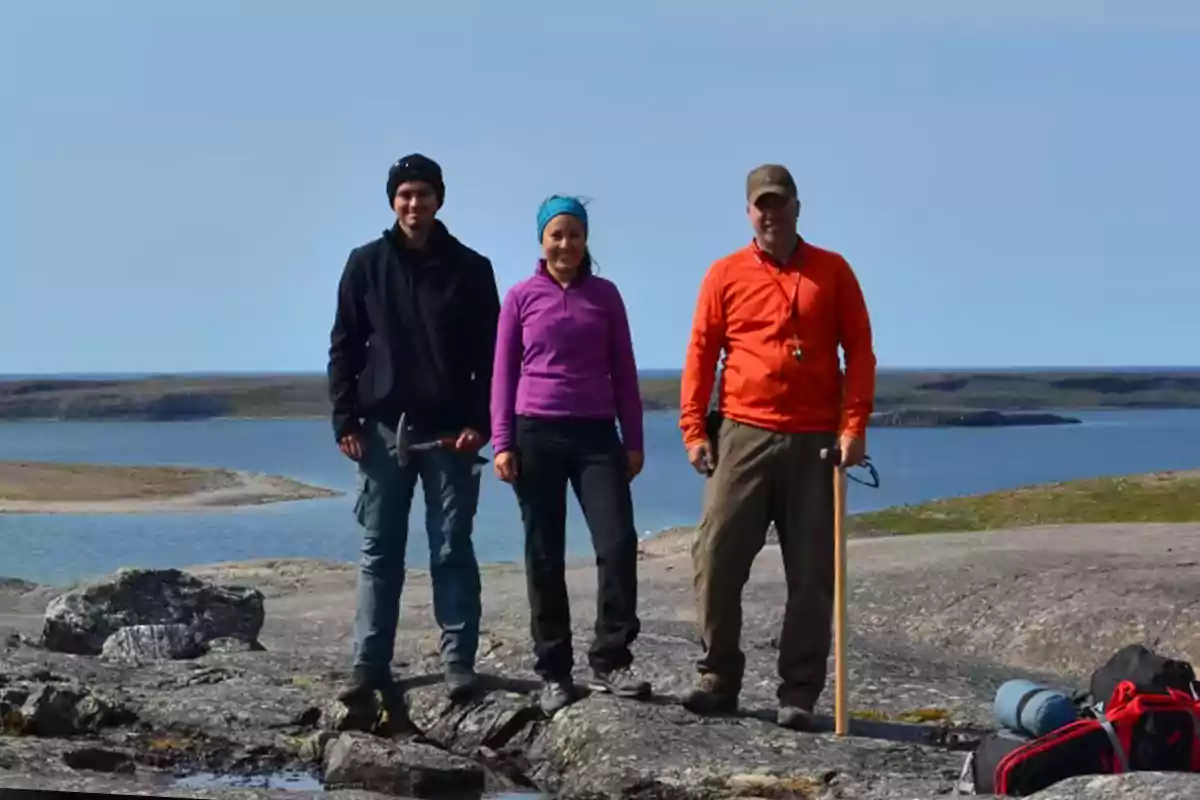 Three people standing on rocks by a lake in a natural landscape, with hiking gear and tools Three people standing on rocks by a lake in a natural landscape, with hiking gear and tools