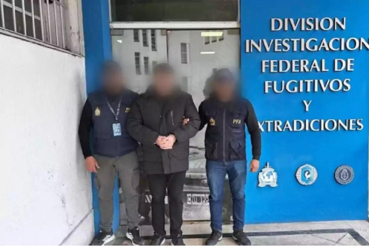 Tres personas con los rostros difuminados posan frente a una pared azul con el texto División Investigación Federal de Fugitivos y Extradiciones
