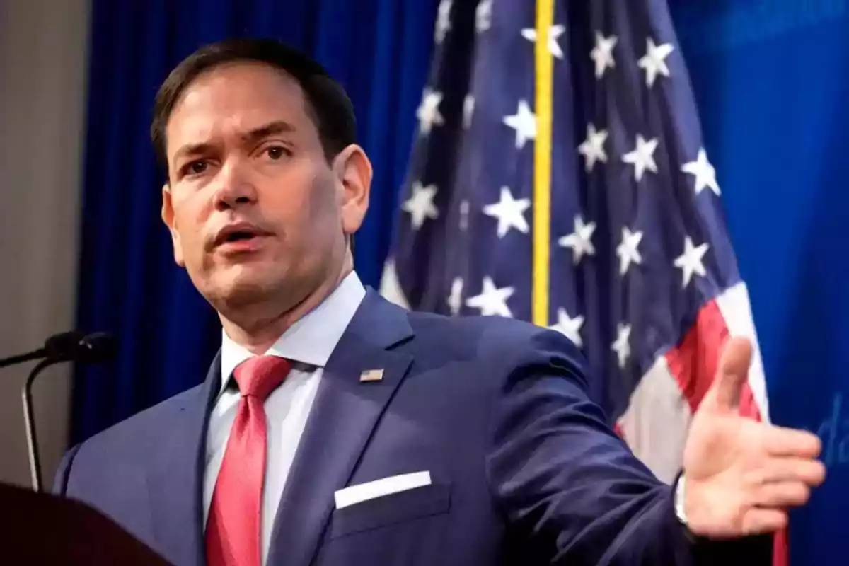 Man in a blue suit and red tie speaking in front of a microphone with a United States flag in the background Man in a blue suit and red tie speaking in front of a microphone with a United States flag in the background