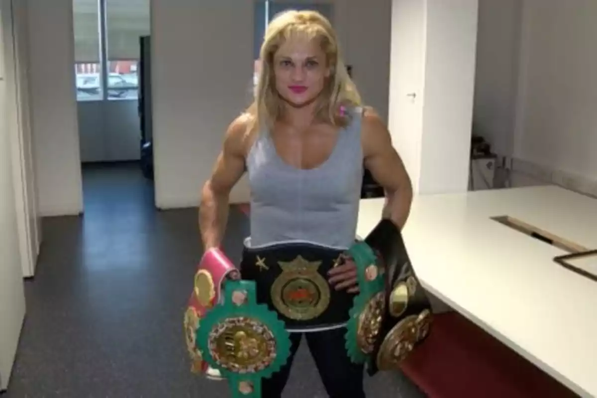 Mujer musculosa con cabello rubio y camiseta gris posando en una oficina mientras sostiene varios cinturones de campeonato de boxeo Mujer musculosa con cabello rubio y camiseta gris posando en una oficina mientras sostiene varios cinturones de campeonato de boxeo