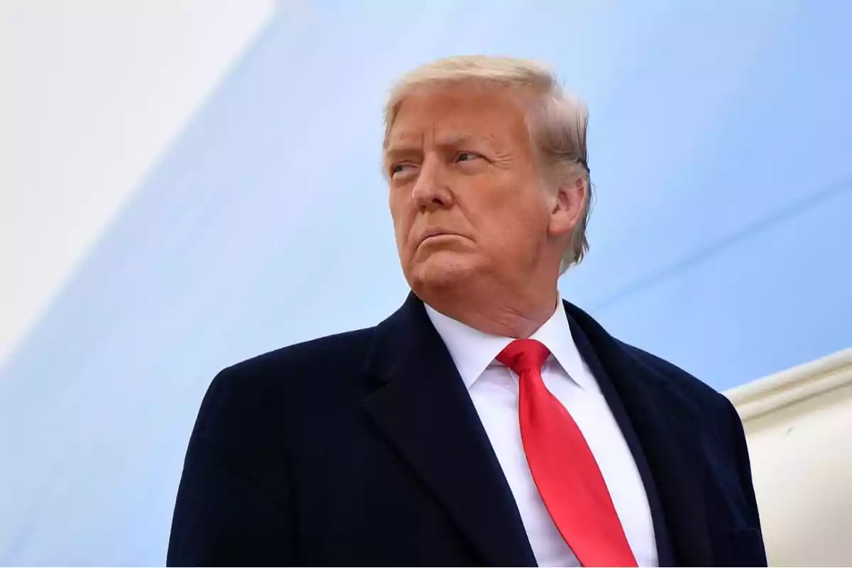Hombre de cabello rubio con traje oscuro y corbata roja mirando hacia la izquierda sobre un fondo azul claro