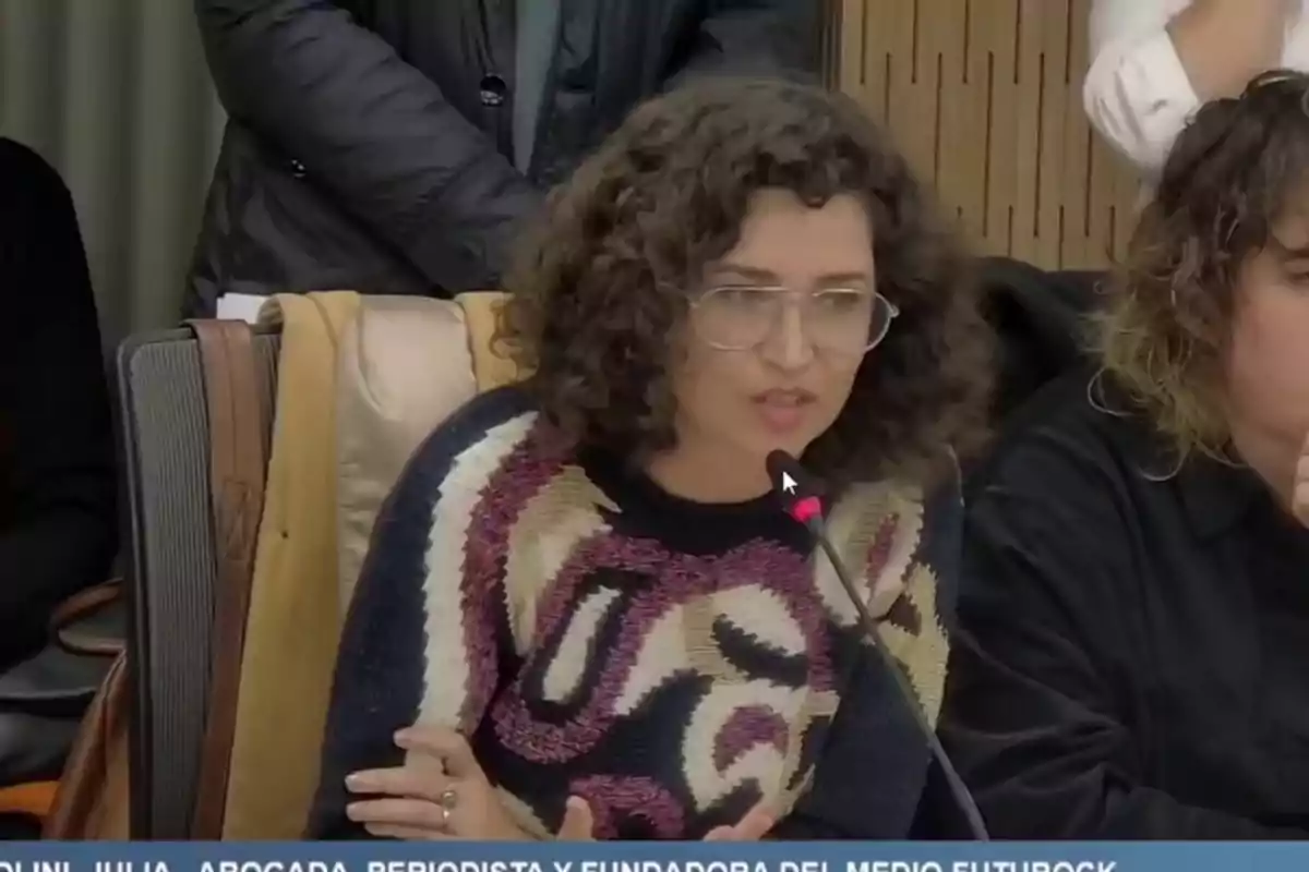 Curly-haired woman with glasses speaking into a microphone at a meeting or conference, seated next to other people Curly-haired woman with glasses speaking into a microphone at a meeting or conference, seated next to other people