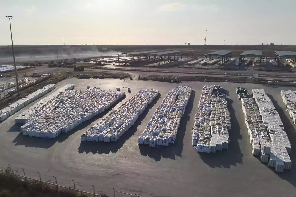Vista aérea de un almacén industrial con grandes filas de materiales apilados en bolsas blancas sobre una superficie asfaltada y estructuras metálicas al fondo Vista aérea de un almacén industrial con grandes filas de materiales apilados en bolsas blancas sobre una superficie asfaltada y estructuras metálicas al fondo