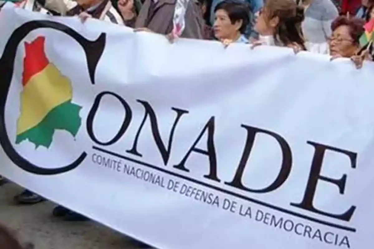 Personas sostienen una pancarta blanca con el logo de CONADE y el texto Comité Nacional de Defensa de la Democracia durante una manifestación