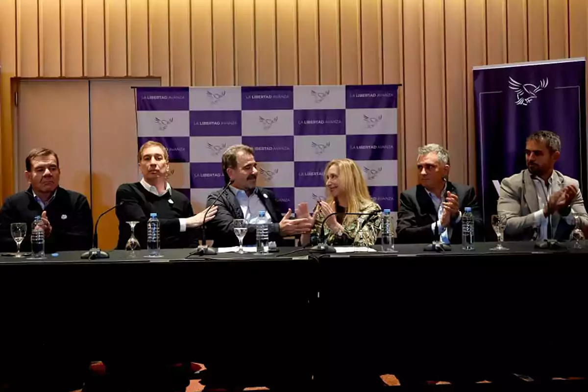 Six people seated at a long table during a conference or political event with La Libertad Avanza banners in the background and water glasses on the table.