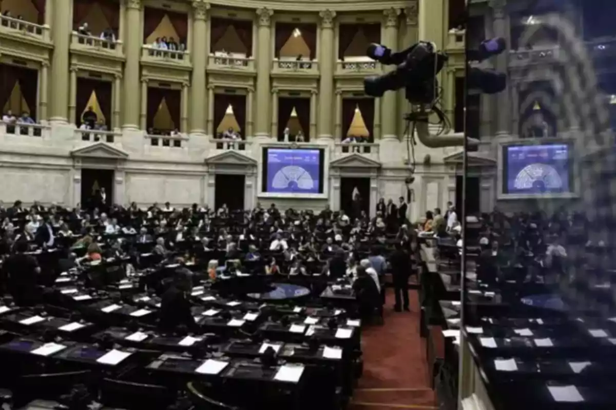 Vista general del recinto de una cámara legislativa con numerosos diputados sentados en sus bancas y una gran pantalla al fondo Vista general del recinto de una cámara legislativa con numerosos diputados sentados en sus bancas y una gran pantalla al fondo