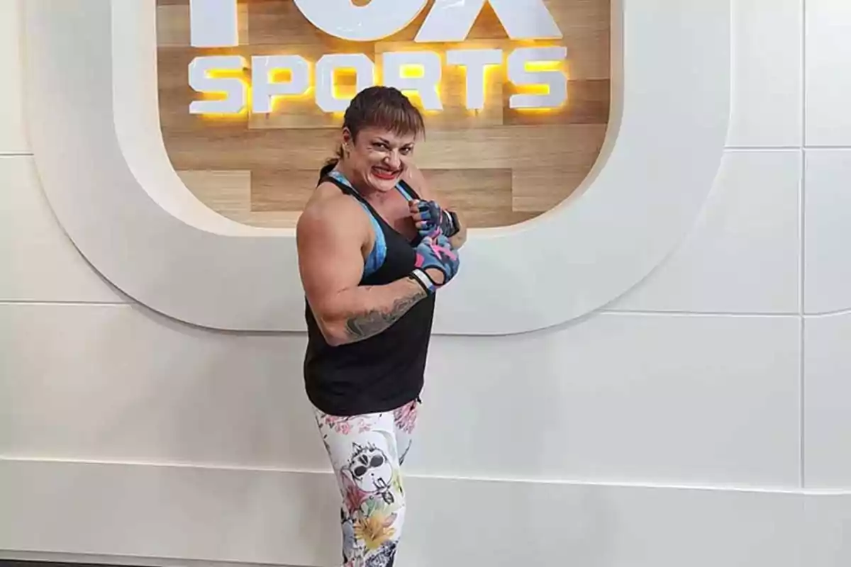 Mujer musculosa posando con guantes deportivos frente a un letrero de Fox Sports Mujer musculosa posando con guantes deportivos frente a un letrero de Fox Sports