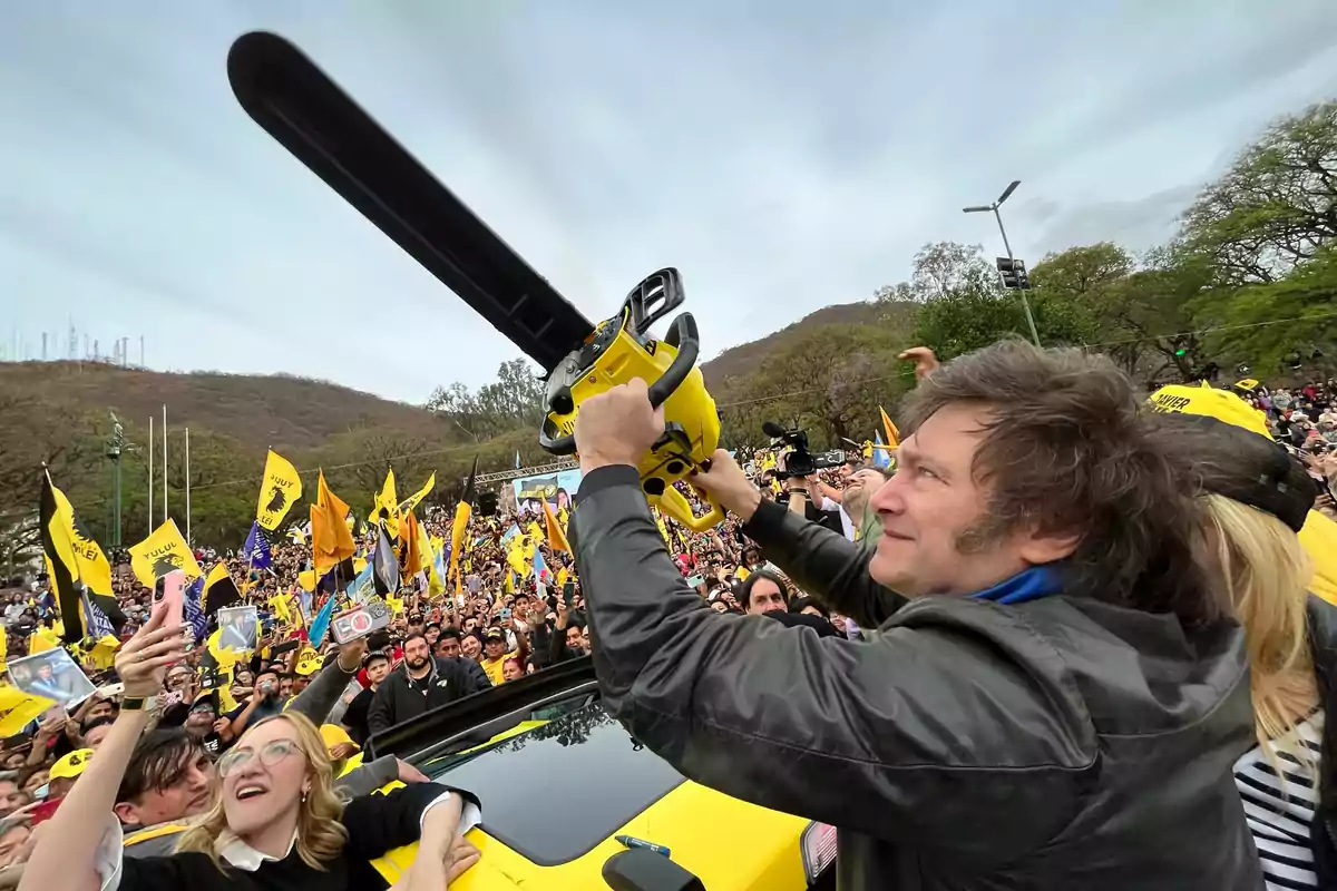 Hombre sonriente sostiene una motosierra amarilla en alto frente a una multitud con banderas amarillas y negras en un evento al aire libre