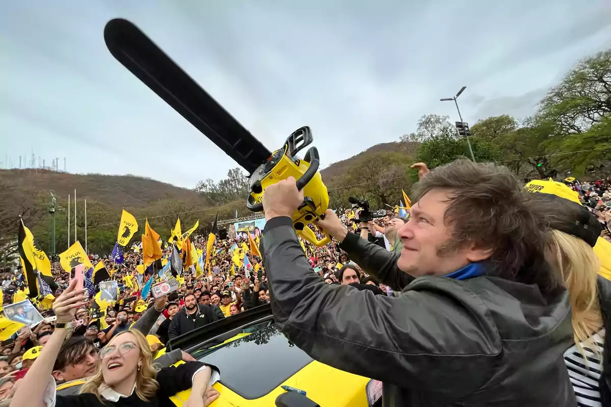 Smiling man holds a yellow chainsaw above his head while a crowd with yellow flags surrounds him at an outdoor event.