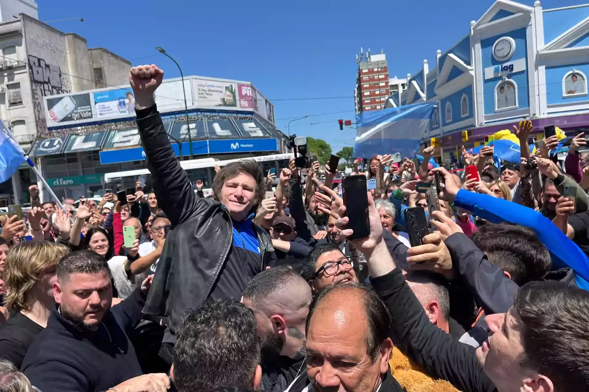Un hombre con chaqueta de cuero levanta el puño mientras una multitud lo rodea y toma fotos con sus teléfonos en una calle de ciudad bajo un cielo despejado. Un hombre con chaqueta de cuero levanta el puño mientras una multitud lo rodea y toma fotos con sus teléfonos en una calle de ciudad bajo un cielo despejado.