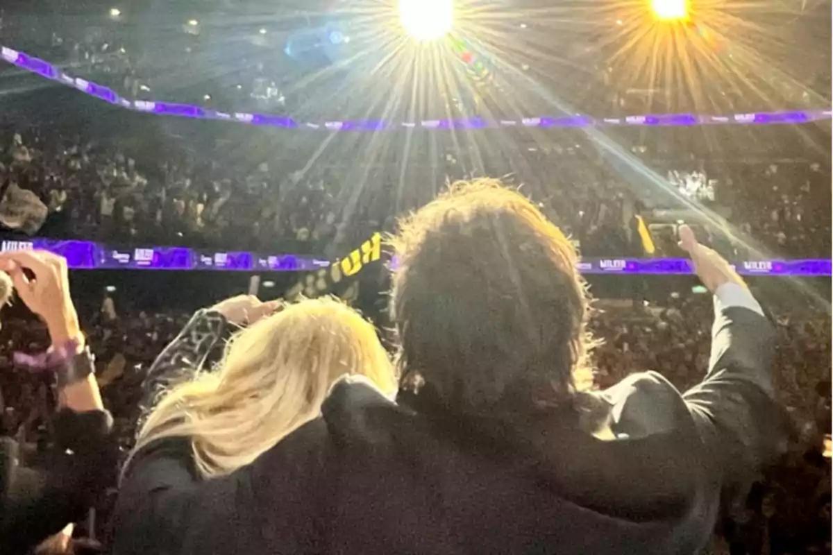 Dos personas de espaldas levantan los brazos frente a una multitud en un estadio iluminado por luces brillantes Dos personas de espaldas levantan los brazos frente a una multitud en un estadio iluminado por luces brillantes