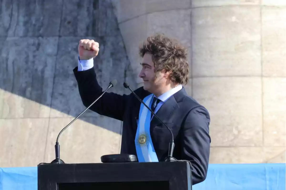 Hombre con banda presidencial argentina levantando el puño derecho mientras da un discurso en un podio al aire libre