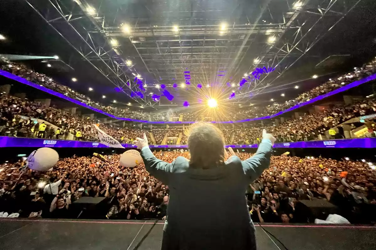 Persona de espaldas en el escenario de un auditorio lleno de gente con los brazos levantados mientras el público aplaude y sostiene globos y pancartas