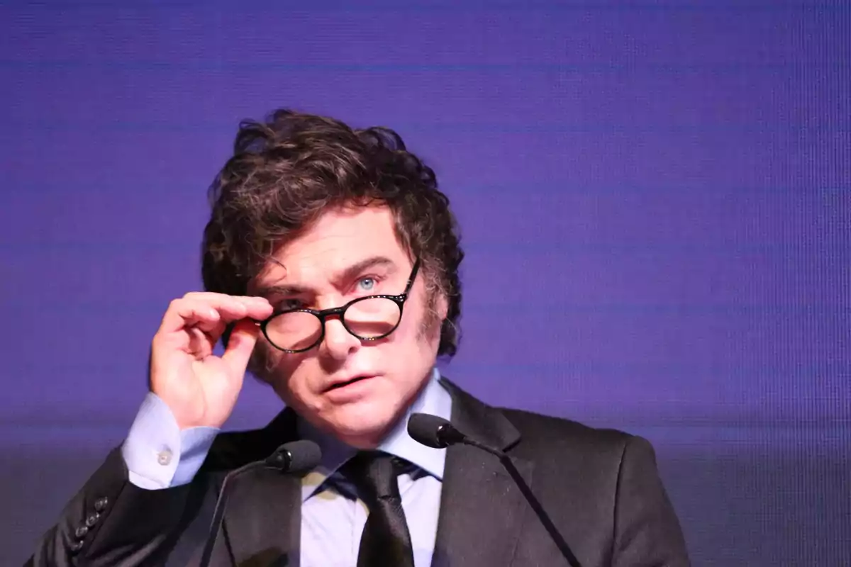 Dark-haired man with glasses and a suit speaking in front of two microphones with a purple background